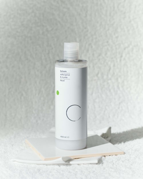 A white bottle with minimal text and design stands on a surface, with a shadow and textured background.