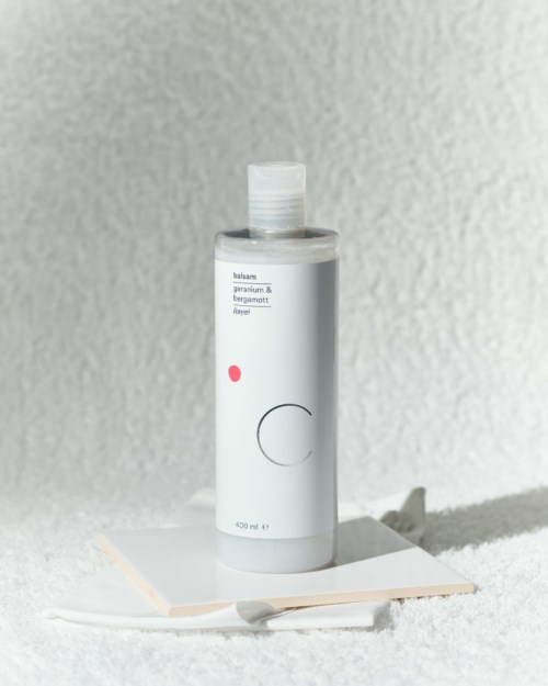 En hvit flaske med enkel design står på en hvit overflate, med en grå, teksturert vegg i bakgrunnen.