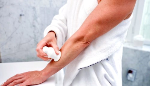 En person påfører creme eller lotion på armen med en hvid håndklæde, i et lyst badeværelse.
