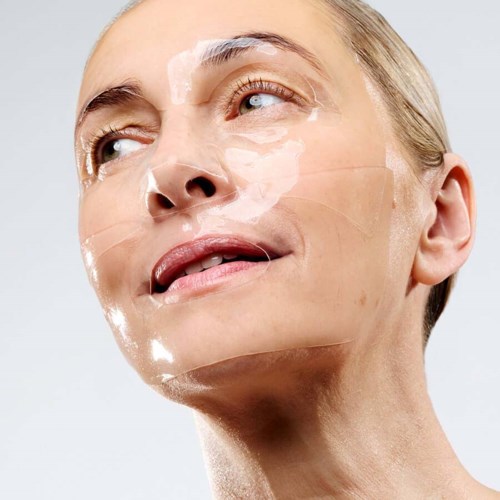 A woman with clear skin, mild makeup, and a transparent facial mask, looking slightly upwards with a gentle smile.