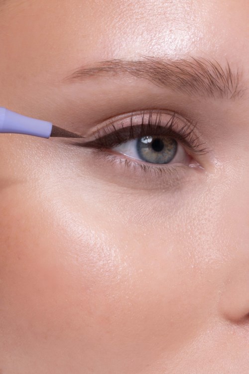 A person applies black eyeliner to their upper eyelid with a small brush.