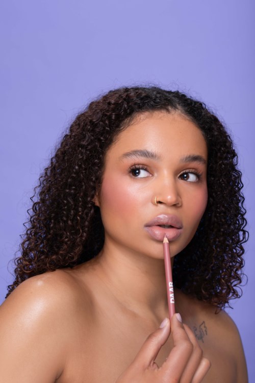 Een vrouw met krullend haar houdt een lippenpotlood dichtbij haar lippen op een paarse achtergrond.