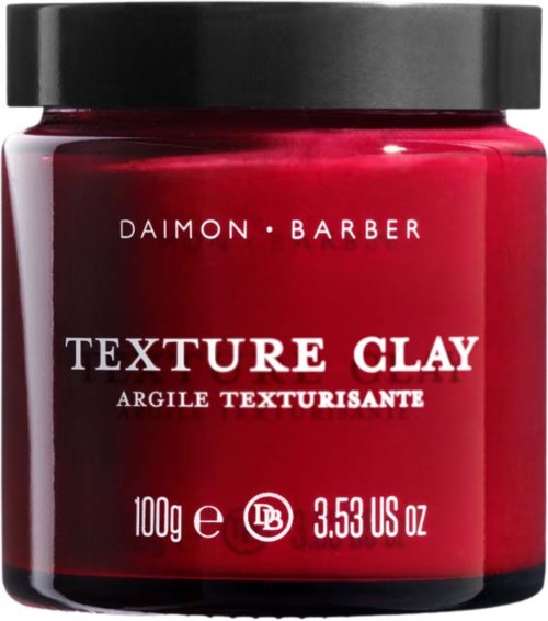 Ein roter Behälter mit schwarzem Deckel, beschriftet 'Daimon Barber Texture Clay' für Haarstyling.