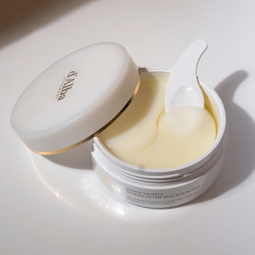 Ein weißer Augencreme-Topf mit offenem Deckel, der Creme und einen Spatel zeigt.