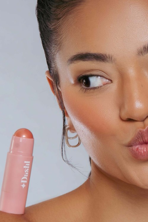 A woman holds a pink deodorant stick, with a playful expression and puckered lips, looking to the side.