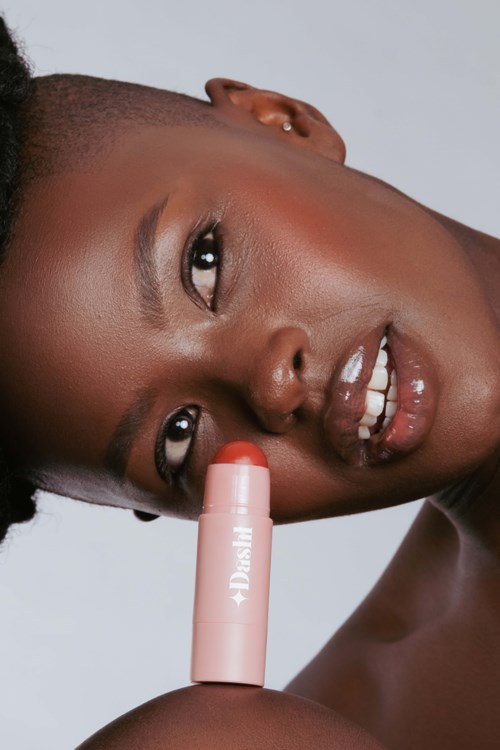 A woman with dark skin holds a lipstick close to her face. She has a big smile and is looking at the camera.