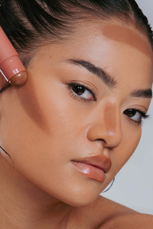 A woman with clear skin, dark hair tied back, and makeup, holding a brown bottle to her face.