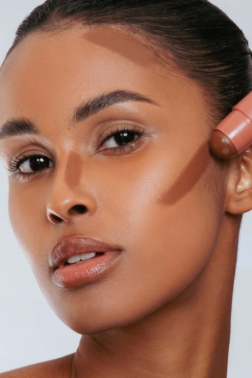 A woman is using a stick makeup product near her face, with smooth skin and well-groomed eyebrows.