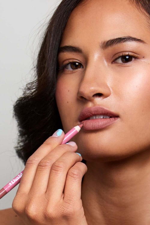A woman applies lipstick with her finger, looking calm with natural makeup and dark hair. The background is plain.
