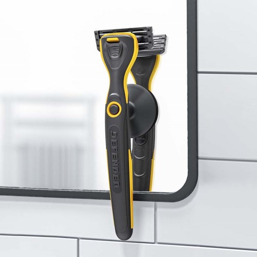 A yellow and black razor is stuck on a mirror in a bathroom. The mirror is attached to white tiled wall.