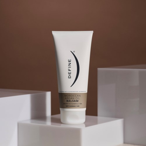 A white tube labeled 'DEFINE Moroccan Argan Oil Balsam' stands on a white block against a brown background.