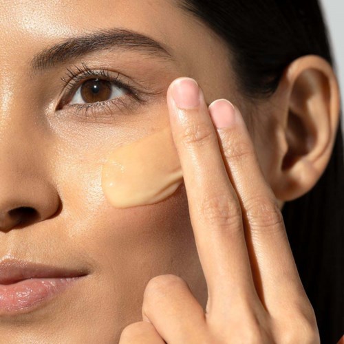 A woman applies foundation on her cheek with her fingers, close-up shot showing her eye and skin coverage.