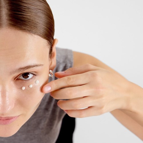 Eine Frau trägt Augencreme auf, kleine weiße Mengen unter dem Auge, während ihre Hand das Gesicht berührt.