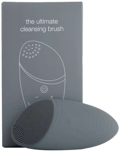 En grå rengöringsborste med rund form och strömknapp, vilar på en låda märkt 'the ultimate cleansing brush.'