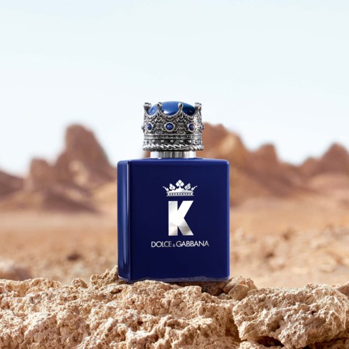 En blå parfymeflaske med et kronehode, plassert på ørkenstein med en fjern fjellandskap i bakgrunnen.