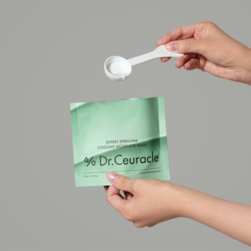 Eine Person hält eine grüne Gesichtsmaske und löffelt weiße Creme oder Gel aus einem kleinen Behälter.