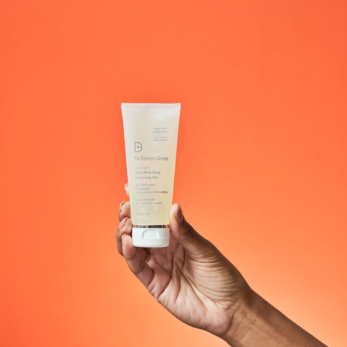 A hand holds a white tube of facial cleansing gel against an orange background.