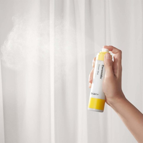 A hand sprays water from a white and yellow spray bottle towards a light-colored surface.