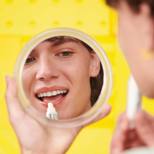 A person looks into a mirror, applying lip gloss with a joyful expression, set against a yellow background.