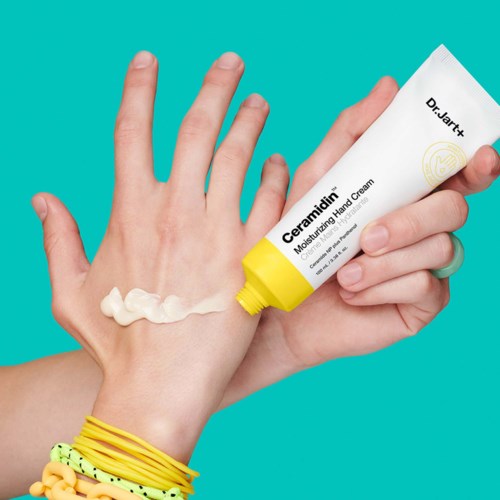 En hånd, der påfører creme fra en tube mærket 'Ceramidien' på bagsiden af en anden hånd, med farverige armbånd på håndleddet.