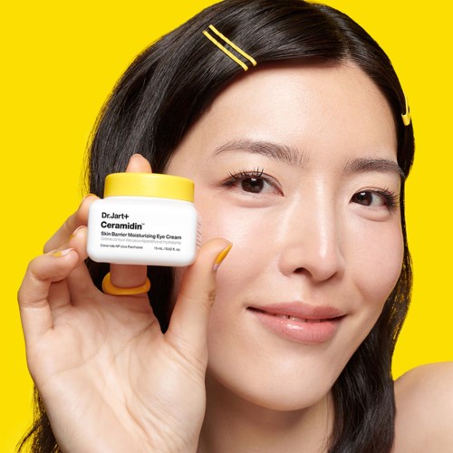 A woman with dark hair holds a small jar of skincare cream. She smiles and wears yellow clips on her hair.