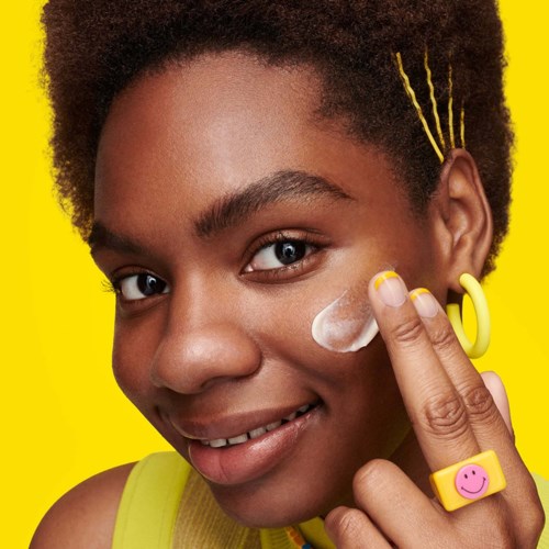 Girl applying white cream to her cheek with colorful jewelry and a bright yellow background.