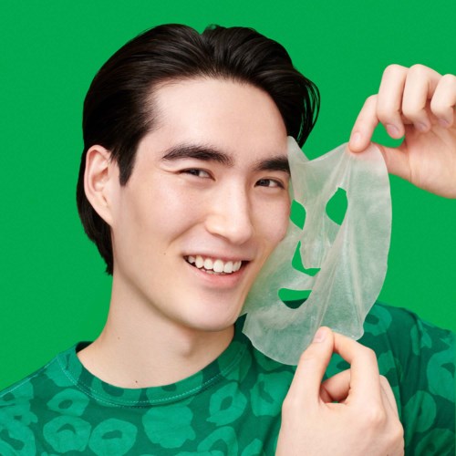 A young man with dark hair holds a translucent sheet with a face cutout in front of a green background and smiles.