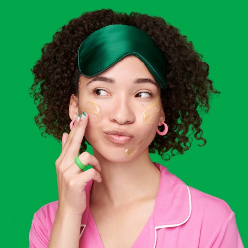 A woman with curly hair applies cream on her face against a green background. She's wearing pink pajamas and a green eye mask.