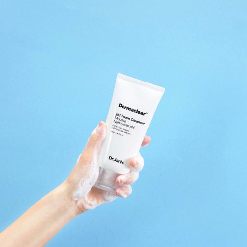 A hand holds a white tube labeled "Dermaclear pH Foam Cleanser" against a light blue background.