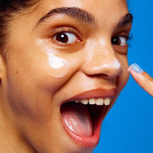 A woman applies cream under her eye, smiling with an open mouth, against a bright blue background.