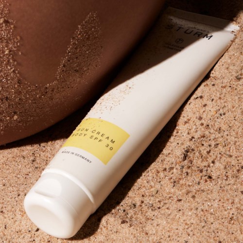 Een witte tube met zonnebrandcrème SPF 30 ligt op zand naast een arm.
