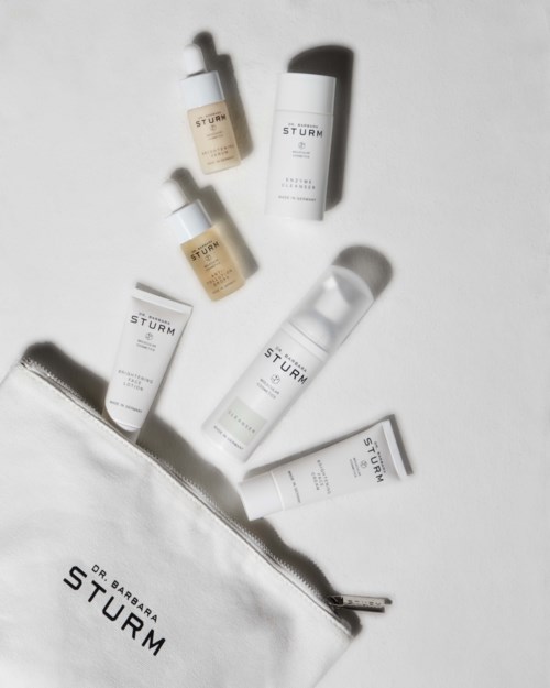 Sju hudpleieprodukter med hvit og beige emballasje og en hvit veske med svart tekst.
