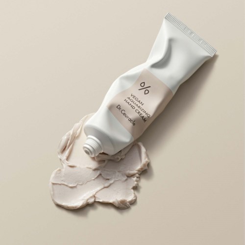 Hvit tube med vegansk hånd- og kroppskrem med beige etikett, sølt på en lys beige overflate.