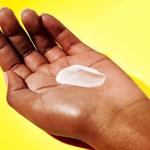A hand holds a white lotion or cream on a bright yellow background.