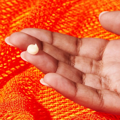 En hånd holder en liten mengde krem eller lotion med en oransje teksturert bakgrunn.