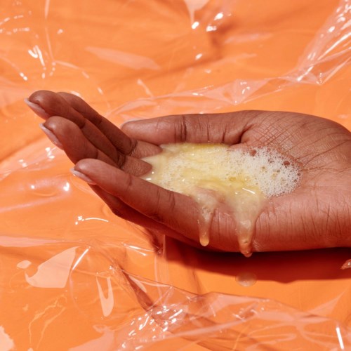 En hand pressar ut en gulaktig vätska i en genomskinlig plastpåse på en orange yta.