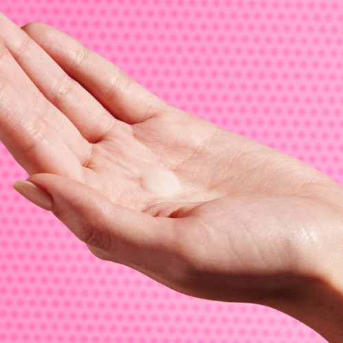 En hånd med litt lotion på en rosa prikkete bakgrunn.