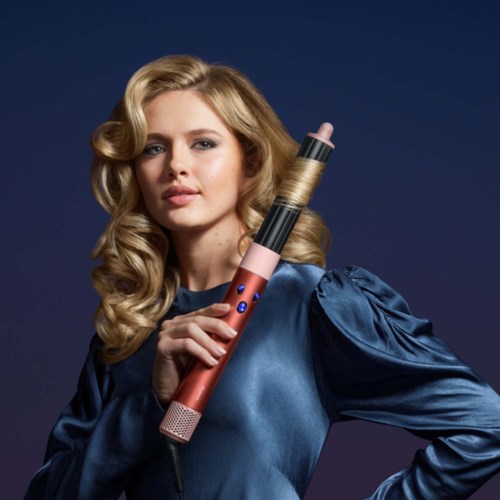 A woman with long, wavy blonde hair holds a pink curling iron and wears a shiny blue blouse.