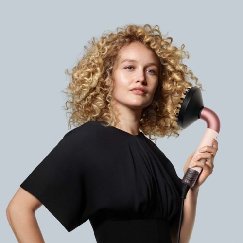 Nainen, jolla curly blonde hair, pitää hiusharjavaa föntistä. Hänellä on musta vaate ja hän katsoo itsevarmasti.