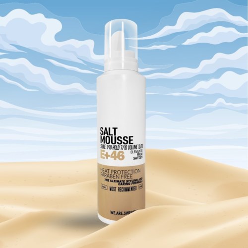 En hvit sprayflaske merket 'Salt Mousse' står på sand med blå himmel og skyer i bakgrunnen.