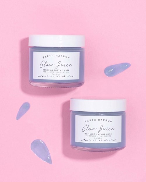 Två burkar med Earth Harbor Glow Juice utvinnande enzymmaska på rosa bakgrund, med lila krämprov runt.