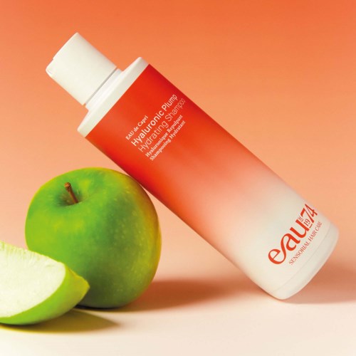 A skincare product bottle lying beside a whole green apple and a sliced apple. The bottle is orange and white.