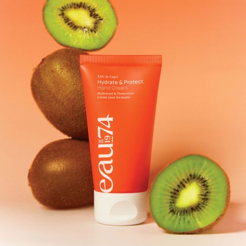 A red hand cream tube with kiwi fruits around it, two whole and two sliced, on a peach background.