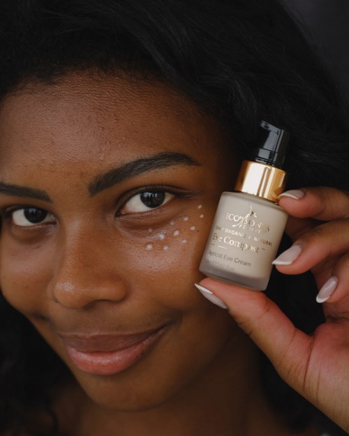 A woman with dark hair and skin applies skincare cream to her face, smiling softly at the camera.