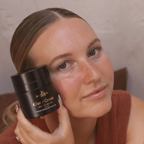 Eine Frau mit Sommersprossen hält einen schwarzen Behälter mit der Aufschrift Glory Cream in der Nähe ihres Gesichts.