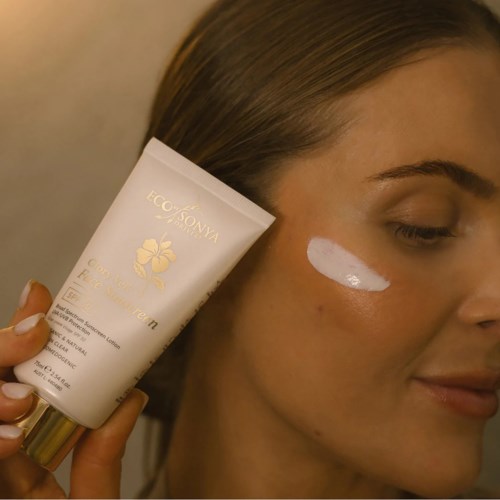 Eine Frau trägt weiße Creme im Gesicht auf, hält eine Tube mit gold- und weißen Design, lächelt leicht.