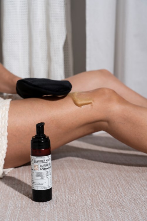 En person påfører lotion eller creme på benet med en pumpflaske i nærheden, i et hyggeligt miljø.