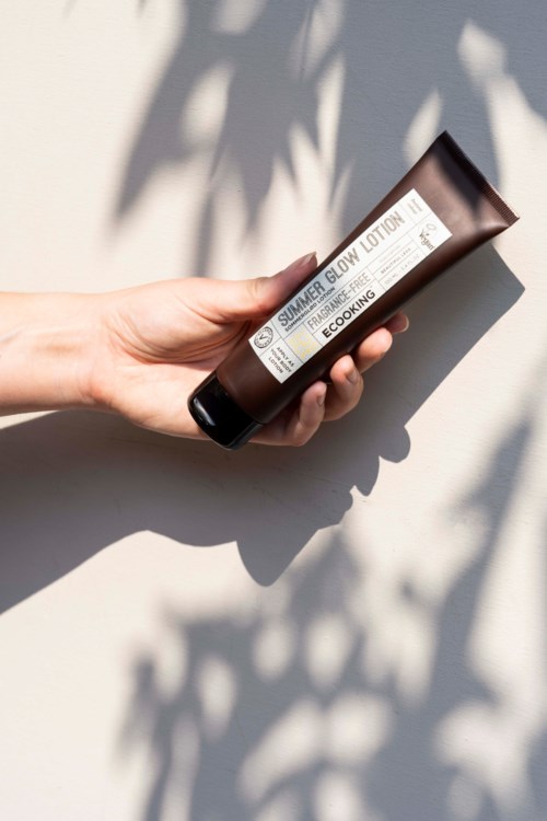 A hand holding a brown tube of summer glow lotion with shadow patterns on the background.