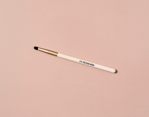 A white brush labeled "EYE PRECISION BRUSH" with a black tip, lying on a pink surface.