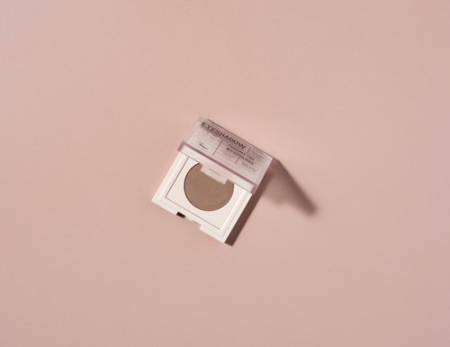 En makeupkompakt med puder inuti är öppen på en rosa yta och kastar skugga.